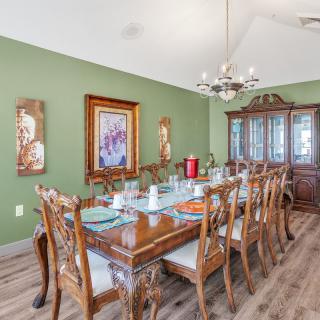 A classic dining room with a large wooden table set for a meal, surrounded by ornate chairs and illuminated by a chandelier. The room features green walls adorned with wall art and decorative plates, as well as a wooden cabinet in the background.
