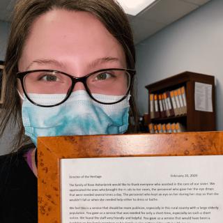 A person wearing glasses and a mask holds a framed document in an office setting.