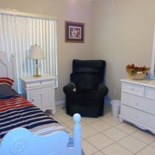 A cozy bedroom featuring a bed with striped bedding, a white nightstand with a lamp, and a comfortable armchair next to a dresser.