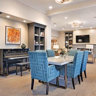 A modern living and dining area featuring blue upholstered chairs, a piano, contemporary artwork, and built-in shelves.