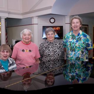 A group of four elderly women smiling while standing behind a piano indoors.