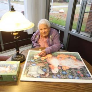 An elderly woman assembling a jigsaw puzzle at a table under the warm light of a lamp.