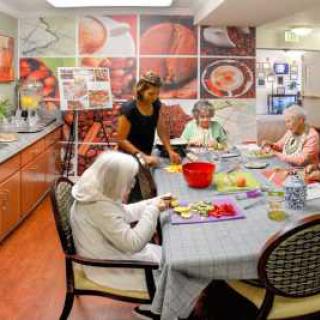 Seniors engaged in an activity with a caregiver in a communal kitchen setting.