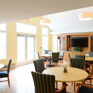 A bright dining area with tables and chairs, featuring large windows and a television.