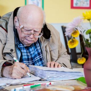 An elderly man deeply focused on writing in a notebook indoors.