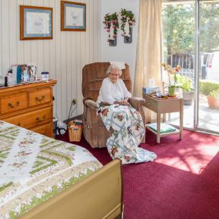 An elderly woman sits comfortably in a cozy bedroom filled with sunlight, surrounded by personal decor and a warm quilt.