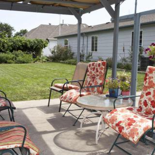 A cozy backyard patio with cushioned chairs and a table under a pergola, overlooking a well-maintained lawn.