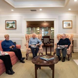 A group of seniors sitting in a cozy living room, reading newspapers and conversing.