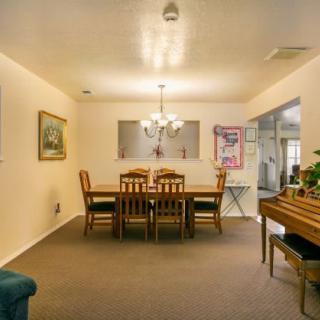 A cozy dining room with a wooden table and chairs, illuminated by a chandelier, featuring artwork on the walls and a piano in the corner.