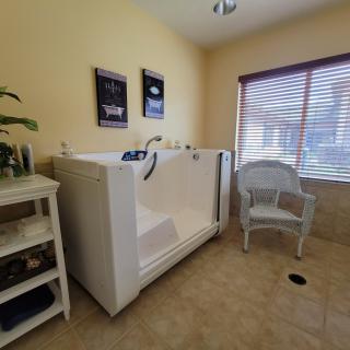 A cozy room featuring a large, accessible bathtub with a nearby chair and shelf, illuminated by natural light from the window.