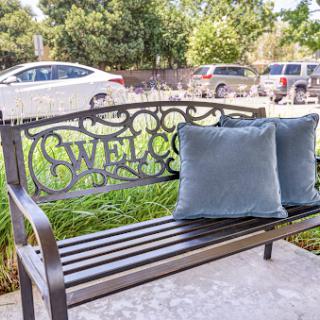 A welcoming bench with decorative pillows, set against a backdrop of greenery and parked cars.