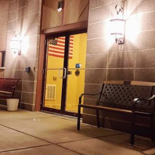 A nighttime scene outside a building entrance with benches and lit lanterns, featuring an American flag visible through the glass doors.