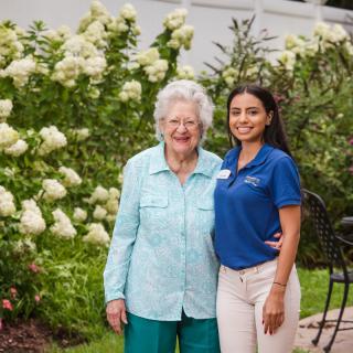 An elderly woman and a younger woman smiling together in a garden full of white flowers.