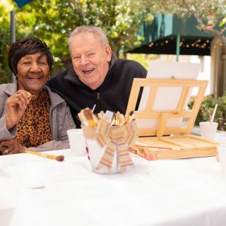 Two seniors enjoying a painting session outdoors, surrounded by art supplies.