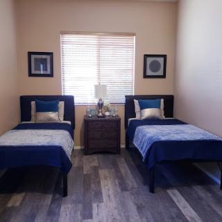 A cozy bedroom featuring twin beds with blue bedding and a nightstand in between, illuminated by natural light from a window.