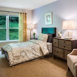 A cozy bedroom with a neatly made bed, side table with lamps, and a large window.