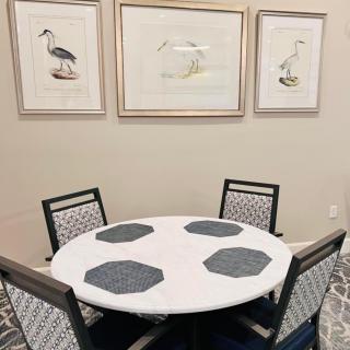 A round dining table with four chairs is set with placemats, and the surrounding wall features framed bird artwork.