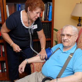 A nurse checks the blood pressure of an elderly man seated in a comfortable chair at home.