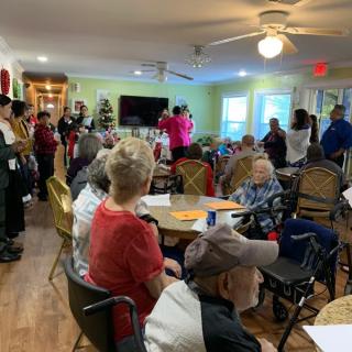 A social gathering of seniors in a community center, with people seated and standing around, engaging in conversations.
