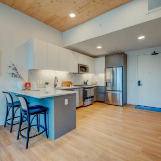 A modern kitchen with stainless steel appliances and a minimalist design, featuring a breakfast bar and wood flooring.