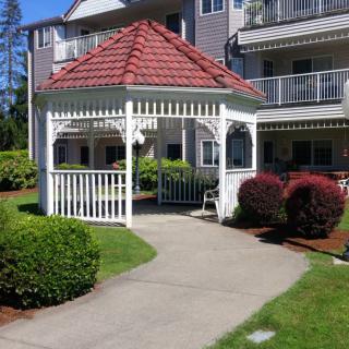 A charming gazebo surrounded by well-maintained gardens and a paved pathway, set in front of a residential building.