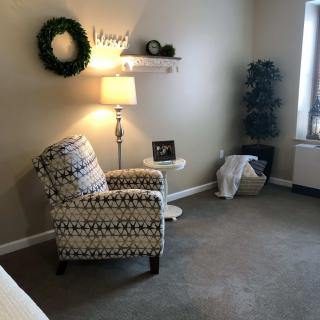 A cozy living room corner with a patterned armchair, floor lamp, decorative wreath, and a small round table.