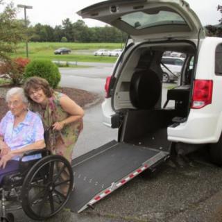 A caregiver assists an elderly person in a wheelchair near a vehicle with an accessibility ramp.