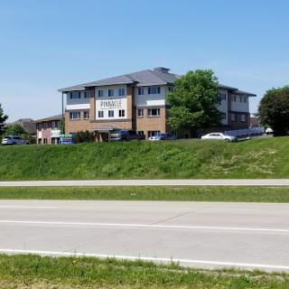A commercial building visible from the roadside, surrounded by greenery under a clear blue sky.