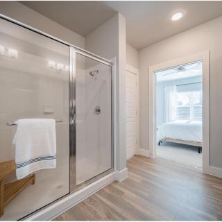 A modern bathroom with a glass shower and wood flooring leading to a bright bedroom.