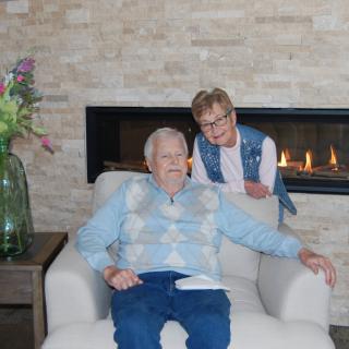 An elderly man sitting in a comfortable chair with a woman standing beside him, indoors by a fireplace.