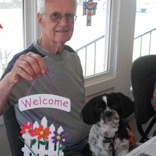 An elderly man sitting indoors with a dog, holding a welcome sign.