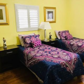 A cozy bedroom with twin beds adorned in colorful bedding, set against sunny yellow walls with artwork and a window featuring shutters.