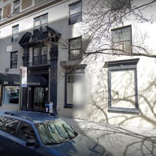 A street view of an urban building with tree shadows on the wall and a parked car nearby.