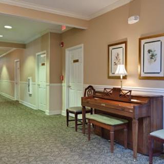 A cozy hallway features a wooden piano accompanied by chairs, framed artwork on the walls, and soft carpeting.