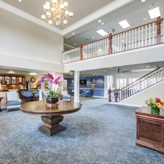 A spacious and elegant lobby with a large chandelier, central table adorned with flowers, carpeted floor, and a staircase leading to an upper level.