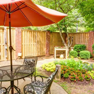 A cozy patio area with a table, chairs, and an umbrella surrounded by a garden.