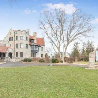 A historic stone mansion with a large lawn and towering trees, set against a clear blue sky.