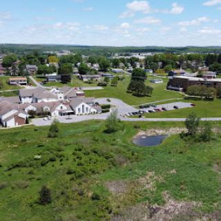 An aerial view of a suburban neighborhood featuring residential buildings, a pond, and lush green spaces under a partly cloudy sky.