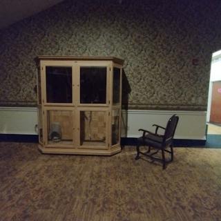A vintage cabinet and a rocking chair sit in an empty room with patterned wallpaper.