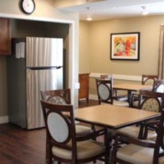 A cozy dining area with tables, chairs, and a refrigerator, featuring warm lighting and artwork on the wall.
