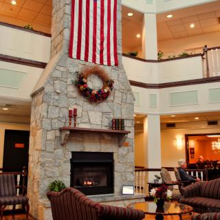 A cozy lobby with a central stone fireplace, adorned with an American flag and a decorative wreath, surrounded by comfortable seating.
