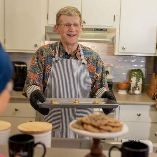 A person wearing an apron and oven mitts holds a tray of freshly baked cookies in a kitchen.