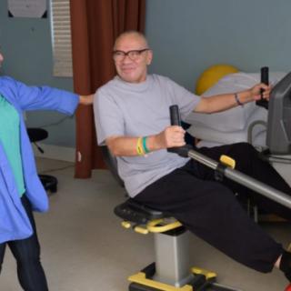 An older adult using a rowing machine with assistance in a gym setting.