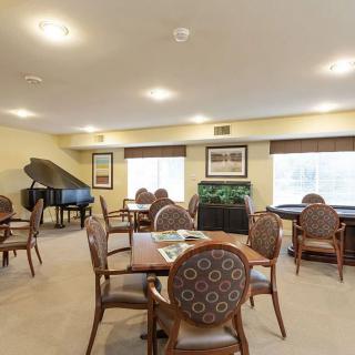 A community room featuring a piano and several tables with chairs, well-lit by natural light through large windows.