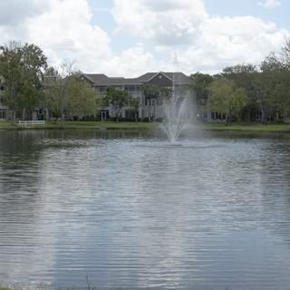 A scenic view of a lake with a fountain, surrounded by trees and buildings under a partly cloudy sky.