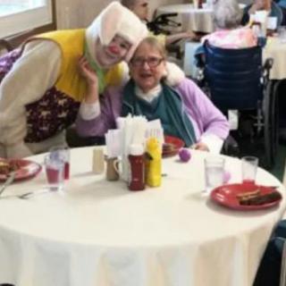A cheerful clown entertains a senior at a nursing home dining area.
