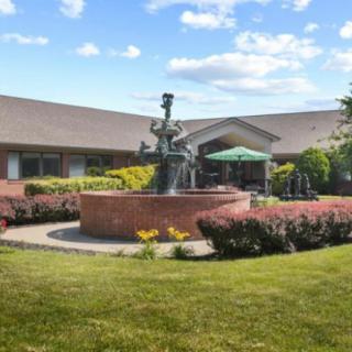 A scenic view of a building with a central fountain surrounded by lush landscaping under a partly cloudy sky.