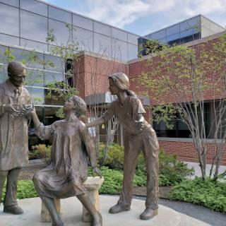 A bronze sculpture depicting healthcare professionals interacting with a patient, set against a modern building backdrop.