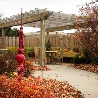 A serene garden with a wooden pergola and a red water pump surrounded by autumn foliage.