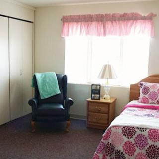 A cozy bedroom featuring a bed with colorful bedding, a nightstand with a lamp, and an armchair by the window.